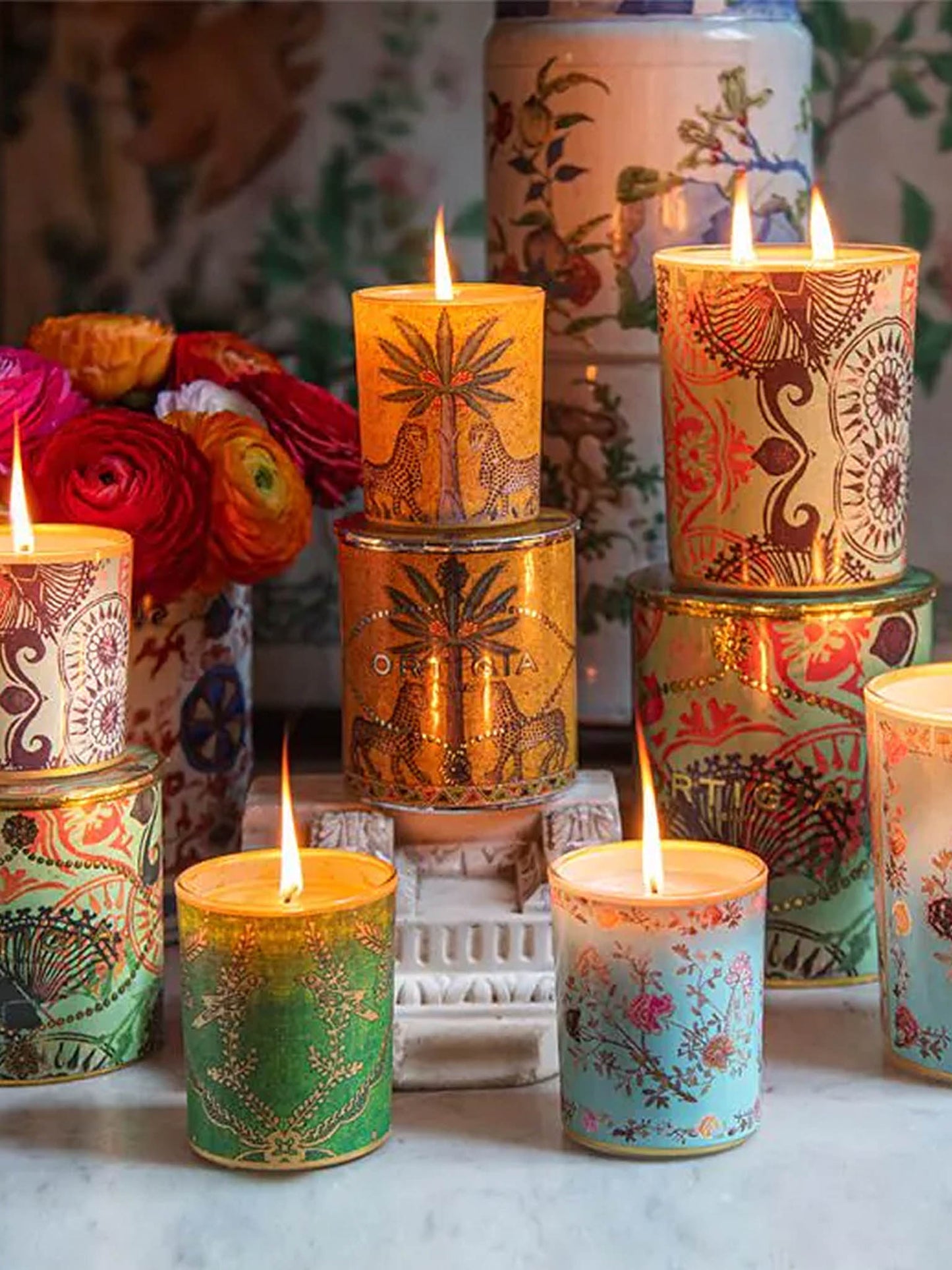 Collection of Ortigia Decorated Candles lit and piles up with their packaging canisters and some flowers